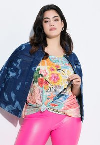 Spijkerjack met een tie-dye patroon, gedragen over een gekleurd graphic t-shirt, gecombineerd met glanzende roze leggings. Opvallende knoopdetail aan de onderkant van het shirt.