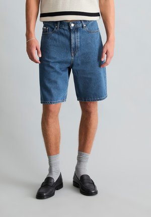 Mann trägt blaue Jeans-Shorts mittlerer Länge, graue Socken, schwarze Loafers und ein cremefarbenes ärmelloses Stricktop mit schwarzem Besatz.