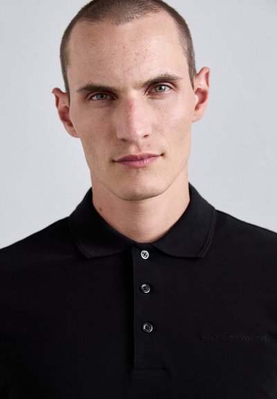 Svart polo-skjorte laget av teksturert stoff, med krage og tre-knapps knapping. Har brodert logo på brystet i en subtil nyanse.