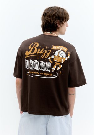 Jeune personne aux cheveux bruns portant un t-shirt marron Buzz Lightyear avec le texte "It's playtime" et "To infinity, and Beyond!" dans le dos.