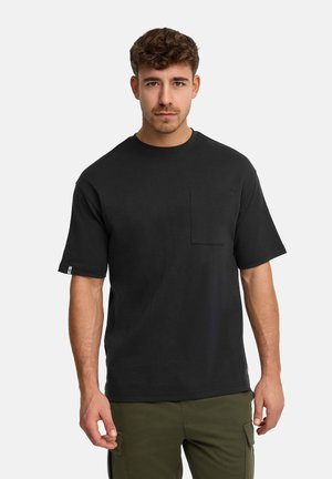 Młody mężczyzna o krótkich kręconych włosach, ubrany w czarny, gładki t-shirt z krótkim rękawem i kieszenią na piersi oraz oliwkowe spodnie typu cargo, stojący nieruchomo.