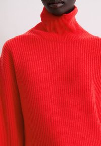 Gerippter Rollkragenpullover in leuchtendem Rot mit hohem Kragen, langen Ärmeln und strukturiertem Strickmuster. Weiches, figurbetontes Material.