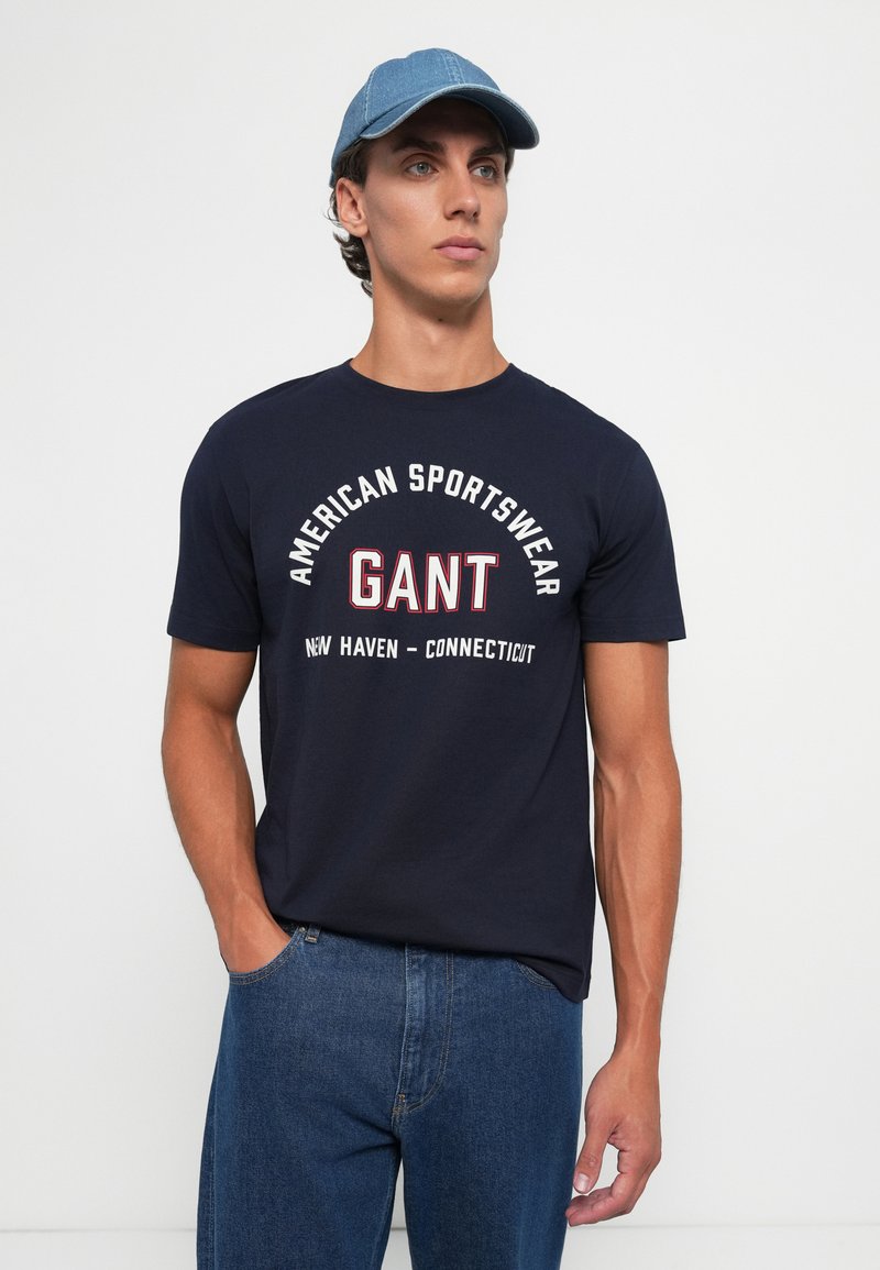 Granatowa koszulka z krótkim rękawem z białym napisem "American Sportswear GANT New Haven - Connecticut." Niebieska czapka noszona tyłem.
