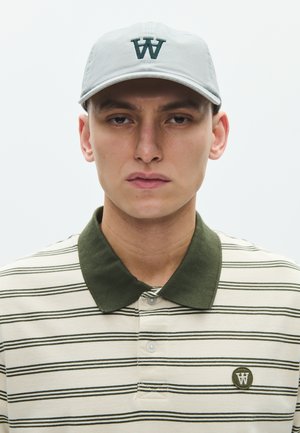 Junger Mann mit hellgrauer Mütze mit dunkelgrünem "W"-Logo und cremefarben gestreiftem Poloshirt mit dunkelgrünem Kragen und passendem Logoaufnäher.