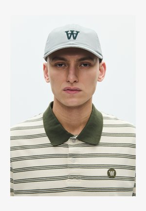 Ung mand iført lysegrå kasket med mørkegrønt "W"-logo og cremefarvet poloshirt med striber, mørkegrøn krave og matchende logo-patch.
