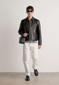 Giacca di pelle nera, camicia a righe, pantaloni bianchi, scarpe nere e una piccola borsa nera. Il modello è in piedi contro uno sfondo semplice.