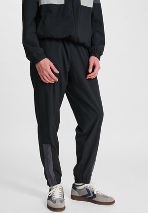 Man wearing black track pants with elastic cuffs, reflective gray side panels, and gray and navy sneakers with white laces.