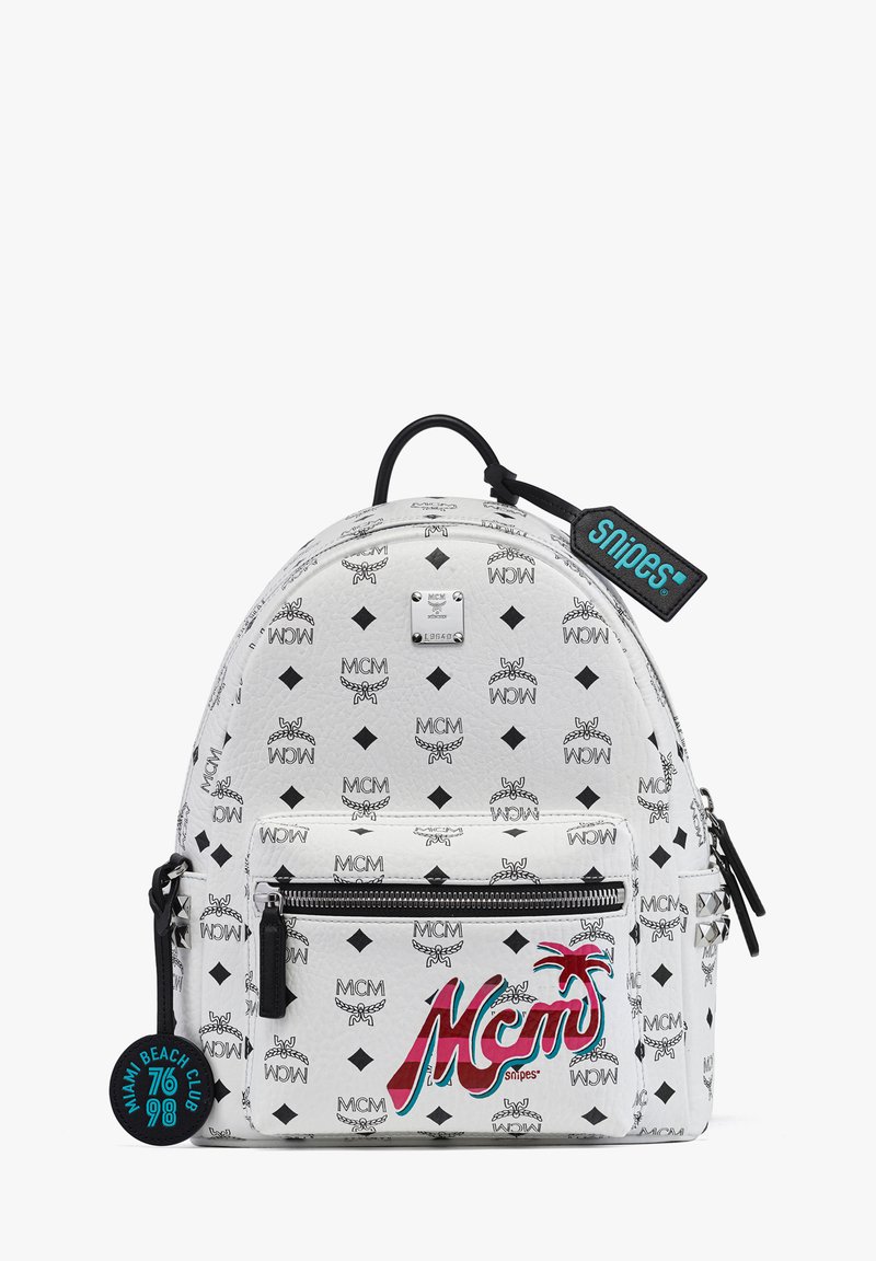 Weißer Rucksack mit MCM-Logo-Muster, runde Form, vorderes Reißverschlussfach und schwarzen Akzenten. Enthält ein dekoratives Etikett mit der Aufschrift "Snipes."
