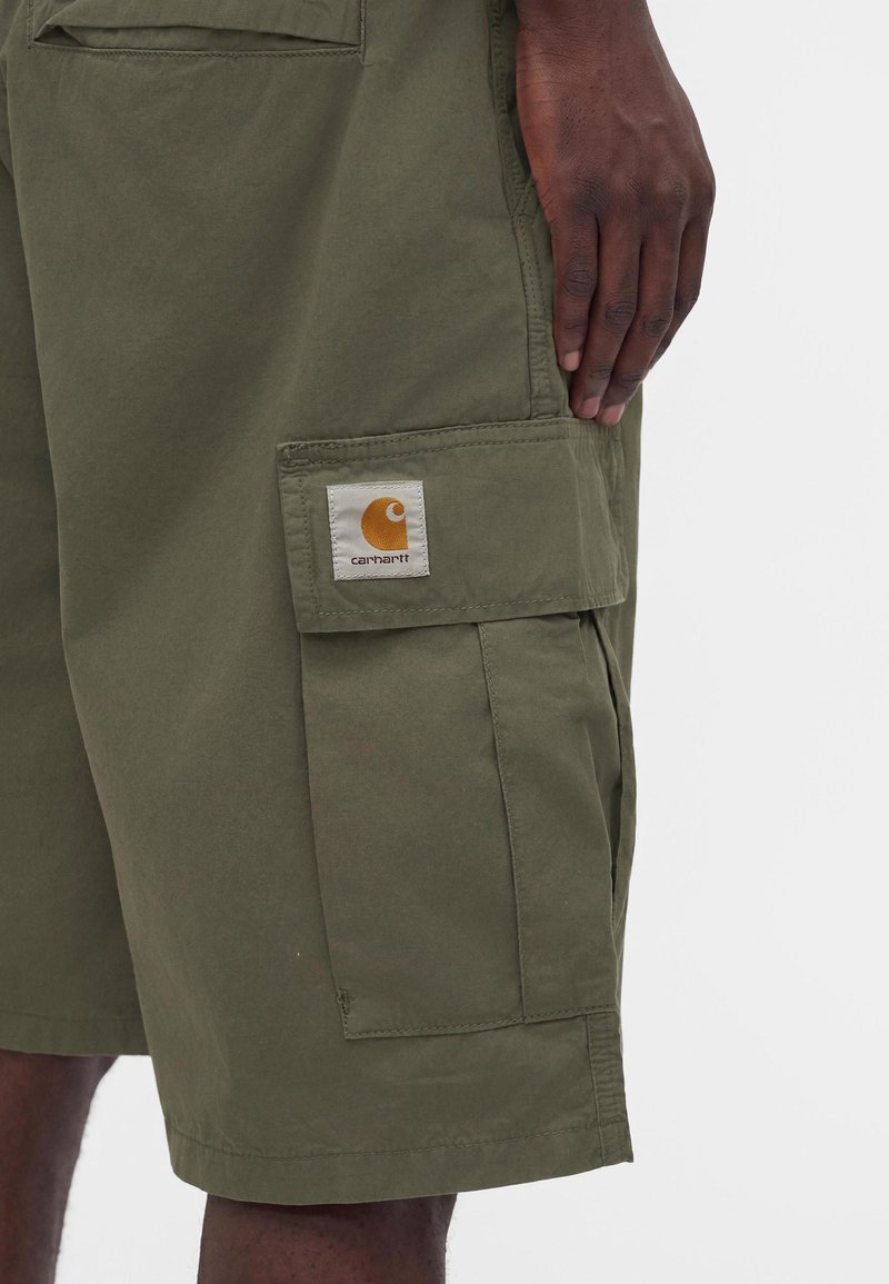 Dunkelhäutige Hand, die auf olivgrünen Cargo-Shorts mit Carhartt-Logo auf der Taschenklappe ruht.