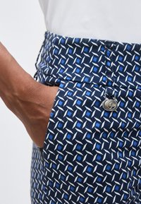 Χέρι στην τσέπη μπλε παντελονιού με γεωμετρικά λευκά και μπλε σχήματα και ασημί κυκλικό κουμπί.