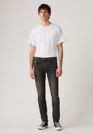Νέος άνδρας στέκεται φορώντας απλό λευκό T-shirt, μαύρο στενό τζιν και μαύρα ψηλά sneakers μπροστά σε ουδέτερο φόντο.