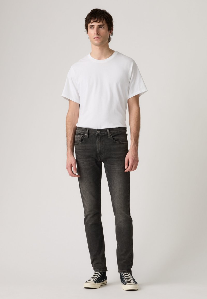 Νέος άνδρας στέκεται φορώντας απλό λευκό T-shirt, μαύρο στενό τζιν και μαύρα ψηλά sneakers μπροστά σε ουδέτερο φόντο.