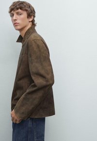 Giacca di camoscio marrone con colletto rialzato, tasche laterali e vestibilità rilassata, abbinata a jeans in denim blu su uno sfondo chiaro.