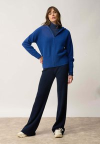 Blå stickad zip-up hoodie med huva, ihopparad med mörkblå ribbade vida byxor. Modell med beige sneakers. Enkel bakgrund.