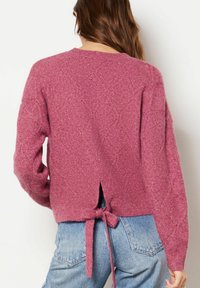 Pull tricoté rose avec motif en losange, dos ouvert noué avec un nœud, ourlet et poignets côtelés, associé à un short en denim bleu.