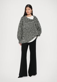 Vrouw met lang zwart haar, gekleed in een grote grijs-witte gebreide trui, witte shirt, zwarte wijde broek, zwarte schoenen en gouden oorbellen.