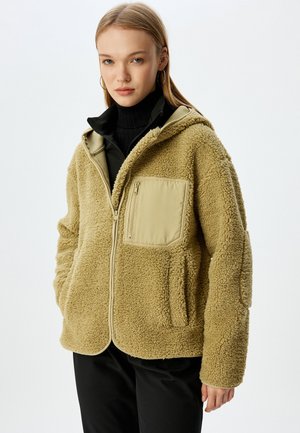 Veste en polaire beige clair avec un zip sur le devant, une capuche et une poche poitrine de couleur beige contrastante. Elle possède un tissu texturé et des poches latérales.