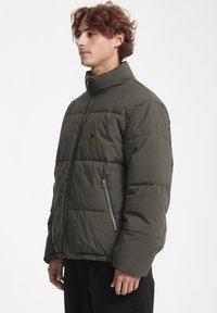 Chaqueta acolchada en verde oliva oscuro con un alto cuello, cierre frontal con cremallera y bolsillos laterales. Presenta un textura acolchada y puños elásticos.
