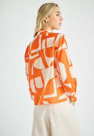 Woman with blonde hair wearing an orange and white geometric patterned long-sleeve shirt and cream pants, facing sideways against a white background.