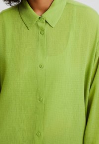 Camicia verde chiaro con bottoni, realizzata in tessuto strutturato, dotata di colletto e sei bottoni abbinati sulla parte anteriore.