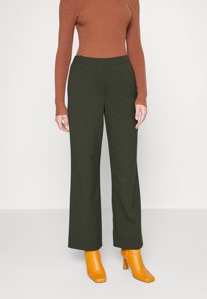 Person wearing dark green straight-leg pants, rust-colored ribbed long-sleeve top, and bright yellow heeled ankle boots, standing on a gray floor.