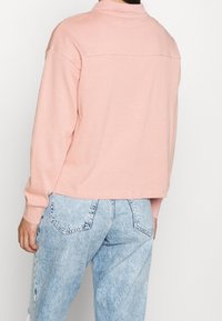 Hellrosa bauchfreier Pullover mit Kragen, langen Ärmeln und einer Nahtdetail über den Rücken, kombiniert mit hoch taillierten blauen Denim-Jeans.