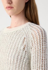 Lichte, crèmekleurige trui met opengehaakt ontwerp, ribbelboord bij de hals en een gestructureerd patroon. Heeft lange mouwen en een ontspannen pasvorm.