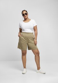 Beige treningsshorts med elastisk midje og sidelommer, kombinert med en hvit t-skjorte og hvite joggesko. Store mørke solbriller er iført.