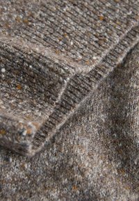 Strukturiertes braunes Strickstoff mit hellen und dunklen Sprenkeln. Verfügt über eine gefaltete Kante und ein gleichmäßiges Rippenmuster im gesamten Material.