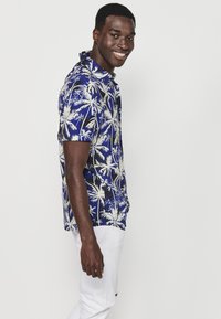 Camicia a maniche corte di colore blu con motivo di palme bianche, design con bottoni e colletto. Indossata con pantaloni bianchi, caratterizzati da una texture liscia.