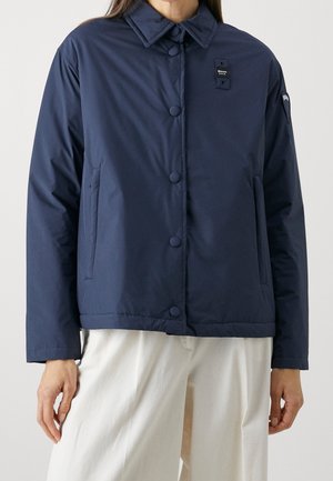 Personne portant une veste boutonnée bleu marine avec un col et un pantalon blanc, debout devant un fond uni.