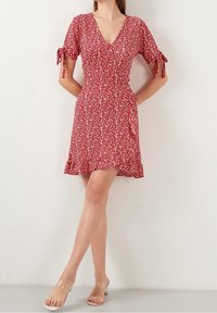Robe fleurie rouge avec un décolleté en V, manches courtes à nouer et ourlet évasé. Confectionnée en tissu léger avec une texture subtile.