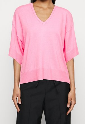 Femme portant un haut rose à manches courtes à col en V et ample, et un pantalon noir, visible du cou aux hanches sur un fond uni.