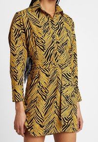 Frau in einem gelb-schwarz gestreiften Hemdkleid mit Gürtel, langen Ärmeln und Kragen, die vor einem einfarbigen Hintergrund steht.