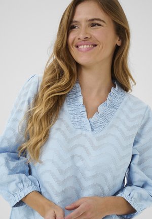 Mlada ženska z dolgimi valovitimi blond lasmi se smeji, oblečena v svetlo modro teksturirano bluzo z volančkastim V-izrezom in nabranimi manšetami.