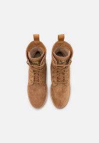 Bottes chelsea en daim marron avec lacets, présentant une texture douce, un bout rond et deux anses sur le col pour un enfilage facile.