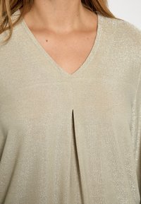 Light beige kreklā ar V veida kakla izgriezumu un īsām piedurknēm, ar vieglu spīdumu un centrālu krokas detaļu, izgatavots no mīkstas, teksturētas auduma.