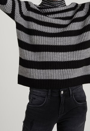 Black and gray horizontally striped knit sweater paired with high-waisted black jeans, cropped above the waist, arms lifted.