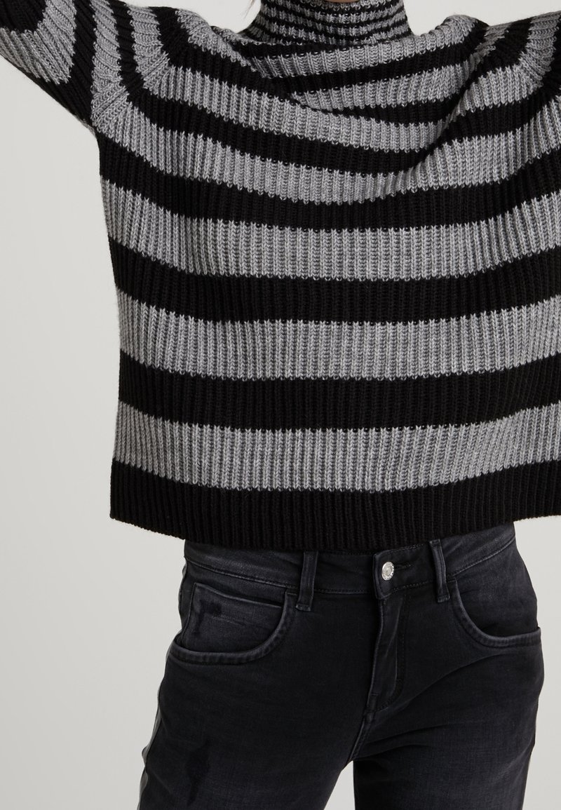 Pull en maille rayé horizontal noir et gris associé à un jean noir taille haute, raccourci au-dessus de la taille, bras levés.