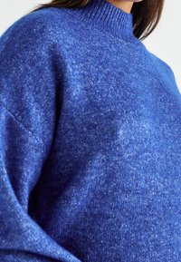 Maglione blu lavorato a maglia con un collo alto, caratterizzato da una consistenza morbida e una vestibilità leggermente aderente, con delle sfumature di blu più chiaro.