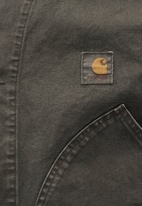 Σκούρο γκρι ύφασμα με υφή, που διαθέτει ετικέτα Carhartt σε τετράγωνη τσέπη με κίτρινο λογότυπο και κόκκινο κείμενο.