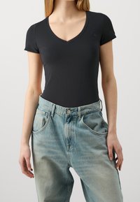 Žena v přiléhavém černém topu s výstřihem do V a krátkými rukávy, zastrčeném do volnějších džínů s vysokým pasem v světlém odstínu, stojící na pozadí bez vzoru.