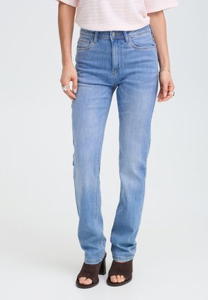 Donna che indossa jeans azzurri a gamba dritta, top rosa pallido a maniche corte con texture e sandali marroni con tacco e punta aperta, in piedi davanti a uno sfondo bianco.