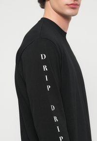 Ung mann som har på seg en svart genser med lange ermer med hvit vertikal tekst "DRIP" gjentatt nedover ermet mot en ensfarget bakgrunn.