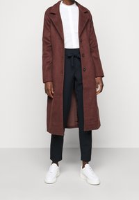 Bordový vlněný kabát dlouhého střihu s klopovým límcem, dvouřadým zapínáním a bočními kapsami. Vytvořeno s černými kalhotami na míru a bílými teniskami.