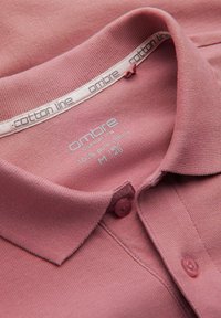 Růžová bavlněná polo košile s texturovaným povrchem, zapínáním na knoflíky a límcem. Etiketa uvádí "100% čistá bavlna, M." Jednoduchý design a barva.