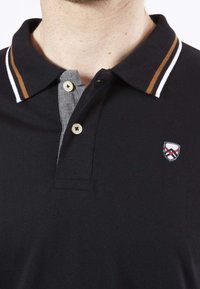 Polo noir avec un plastron gris, deux boutons et un petit blason brodé. Présente des détails rayés en blanc et marron sur le col.