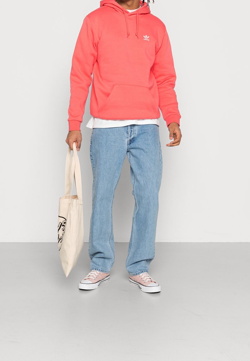 Korallfärgad hoodie med en framficka och logo, kombinerad med ljusblå jeans och rosa sneakers. Tote-väska i handen. Minimalistisk bakgrund.