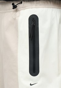 Άσπρα και μπεζ σορτς με κάθετη μαύρη τσέπη με φερμουάρ και μικρό μαύρο λογότυπο Nike κοντά στο κάτω μέρος της τσέπης.