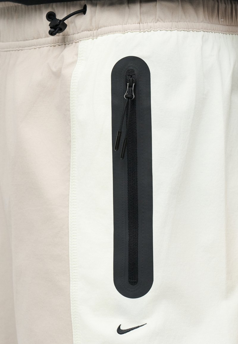 Witte en beige korte broek met een verticale zwarte ritssluitingzak en klein zwart Nike-logo dichtbij de onderkant van de zak.